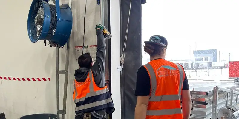 Two technicians in orange safety vests performing garage door tune-ups on a commercial overhead door system.
