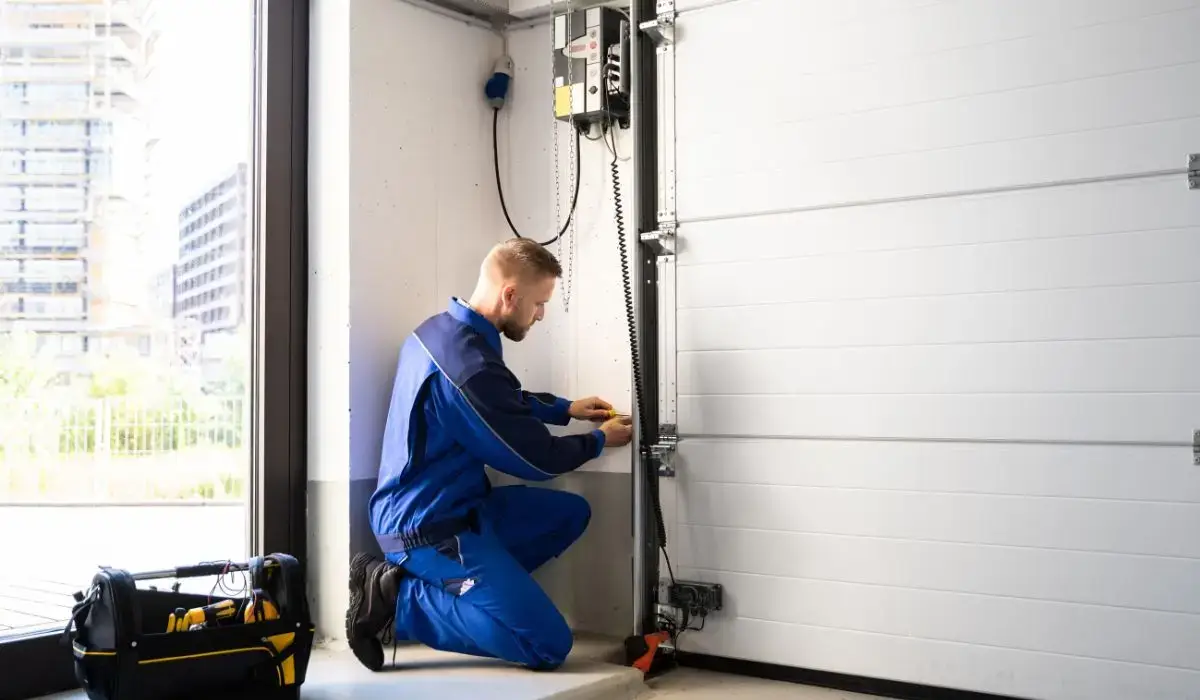 A technician in a blue uniform kneels inside a residential garage while performing garage door tune-ups, adjusting the door mechanism near the wall with tools.